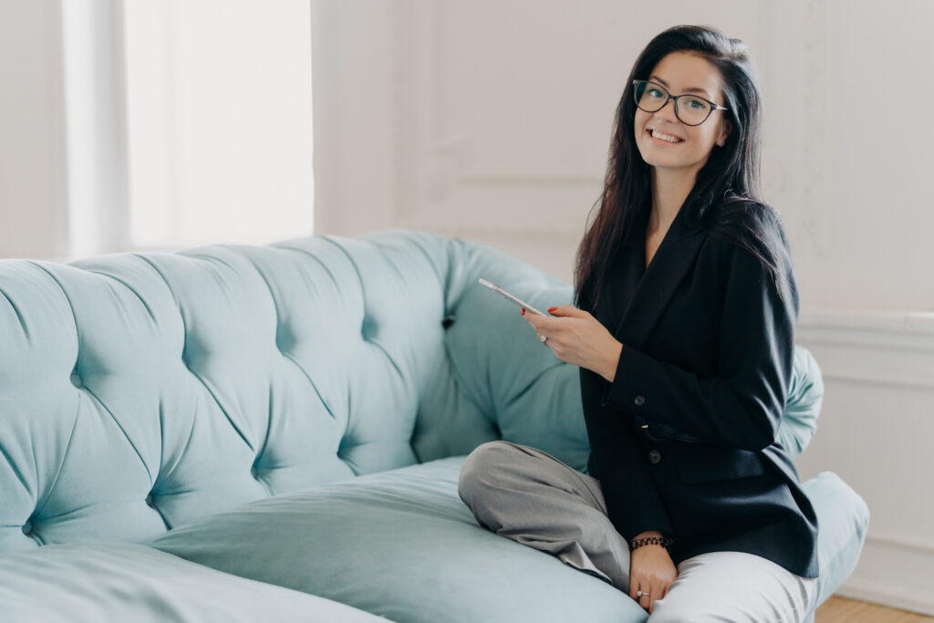 Happy business lady in official clothes, sits on comfortable sofa, uses mobile phone, reads news via network, searches information in cellular, going to phone partner, explores online project